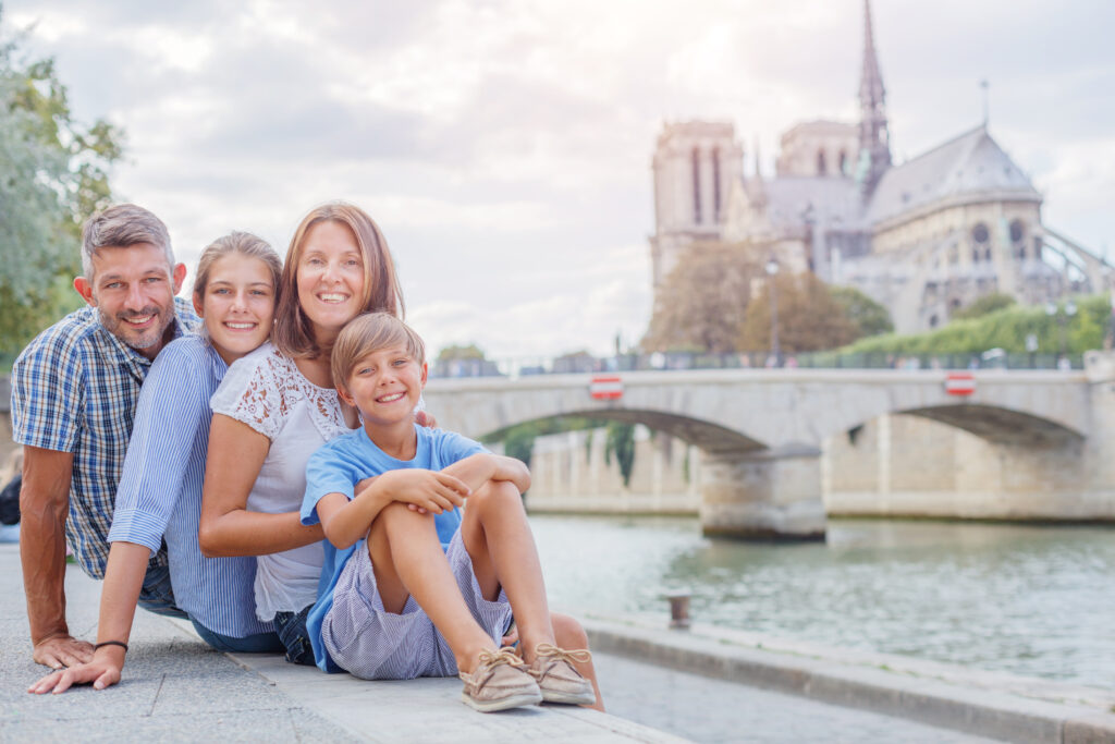 family with parents and two kids sitting near the Seine river with notre dame cathedral behind - visit Paris with kids